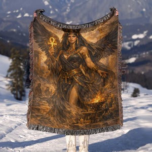 May include: A woven throw blanket featuring a detailed illustration of an Egyptian queen holding an ankh. The artwork is in shades of brown, gold, and black, with fringed edges. The queen has large black wings and is surrounded by flames.