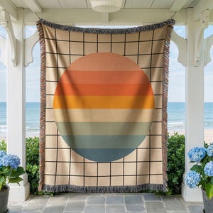 May include: A woven throw blanket featuring a large, centered circle composed of horizontal stripes in shades of orange, yellow, beige, and blue. The background consists of a black grid pattern on a tan base, finished with fringed edges.