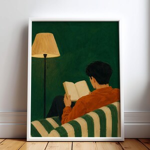 May include: A framed art print depicting a person with dark hair wearing an orange long-sleeved shirt, sitting on a green and white striped chair while reading an open book. A tall floor lamp with a tan shade stands beside them against a dark green wall.