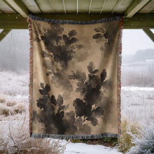 May include: A rectangular woven throw blanket featuring a tan background with a dark brown, watercolor-style botanical leaf pattern. The blanket has a fringed edge along the top and bottom, with a visible texture throughout the woven fabric.