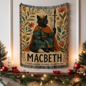 May include: A woven tapestry blanket featuring a black fox wearing a gold crown and a teal and orange royal outfit, holding a spear. The design includes floral motifs and the text MACBETH BY WILLIAM SHAKESPEARE THE THEATER 425 LAFAYETTE STREET.