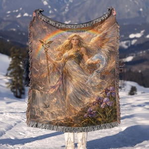 May include: A woven tapestry blanket featuring a celestial woman with wings, holding a staff and a flowing orb. The artwork includes a rainbow, waterfall, and purple irises. The blanket has a fringed edge and is held up against a snowy mountain backdrop.