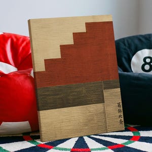 Könnte beinhalten: Ein rechteckiges Wandkunstobjekt aus Holz mit einem geometrischen Design aus horizontalen Streifen in Hellbraun, Dunkelbraun und einer großen, treppenförmigen Fläche in Rostrot. Die Holzmaserung ist sichtbar und schwarze japanische Schriftzeichen sind vertikal aufgedruckt.