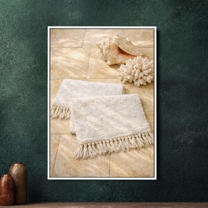 Könnte beinhalten: Zwei cremefarbene Badematten mit Fransen, eine große Muschel und ein Stück Koralle werden auf einer gefliesten Oberfläche präsentiert. Die Matten haben ein strukturiertes Muster. Die Szene ist in einem weißen Rahmen eingefasst.