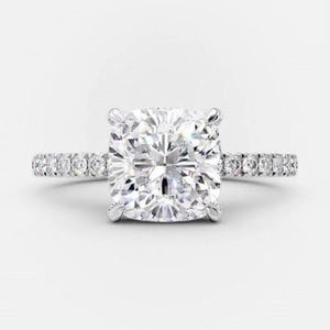 May include: A dazzling engagement ring featuring a large, square-cut diamond as the centerpiece. The band is adorned with smaller, sparkling stones, creating a halo effect around the main diamond. The ring is set in a silver or platinum metal.