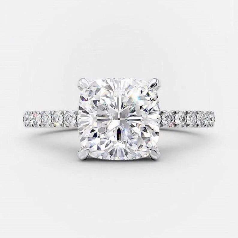 May include: A dazzling engagement ring featuring a large, square-cut diamond as the centerpiece. The band is adorned with smaller, sparkling stones, creating a halo effect around the main diamond. The ring is set in a silver or platinum metal.