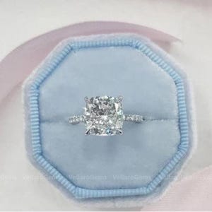 May include: A diamond engagement ring with a large, square-cushion cut center stone. The ring is set in a silver band with smaller diamonds. It rests in a light blue velvet ring box with a pink ribbon.