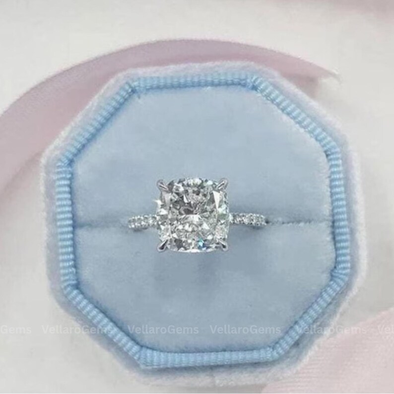 May include: A diamond engagement ring with a large, square-cushion cut center stone. The ring is set in a silver band with smaller diamonds. It rests in a light blue velvet ring box with a pink ribbon.