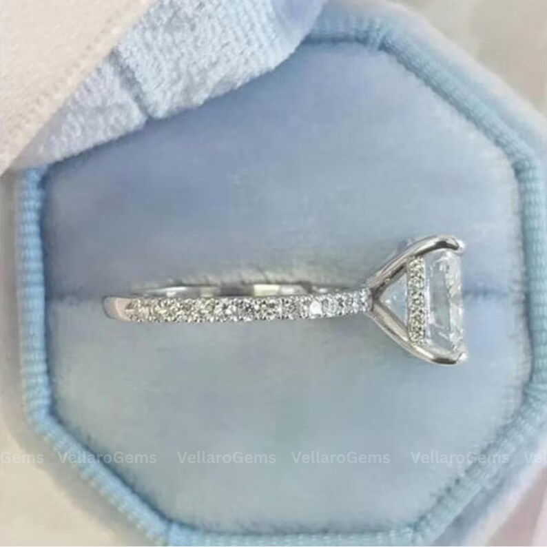 May include: A silver engagement ring with a large, rectangular-cut diamond center stone. The band is adorned with smaller, pave-set diamonds. The ring is presented in a light blue velvet ring box, showcasing the jewelry's sparkle and elegance.