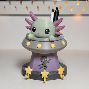 Op de afbeelding: Een bureau-organizer in de vorm van een vliegende schotel met een lichtgroene axolotl die bovenuit steekt. De schotel is grijs met gele ronde accenten en een paarse basis met een donkergrijs alien-silhouet en drie gele stervormige haken.