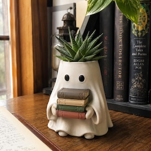 Puede incluir: Una maceta blanca con forma de fantasma y dos ojos negros, que sostiene una pila de cuatro libros pequeños en colores marrón, beige, verde y rojo. Una planta suculenta verde crece en la parte superior de la figura, que tiene un acabado mate.