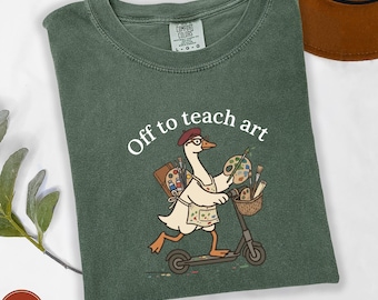 May include: A forest green t-shirt featuring a graphic of a white goose wearing a red beret and glasses, riding a scooter. The goose wears an apron and carries art supplies including a palette and brushes. The text reads, Off to teach art.