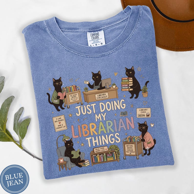 Comfort Colors Blue Jean black cat librarian shirt featuring cute black cats reading books, library checkout desk, book cart, stacked books, library card, cozy reading chair, funny library illustrations, school librarian tee for library staff, reading teacher, book lover and BookTok fans.