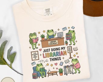 May include: An ivory t-shirt featuring a graphic of green frogs in a library setting. The text reads "JUST DOING MY LIBRARIAN THINGS" with illustrations of frogs reading books, a library checkout desk, a book cart, and various small icons.