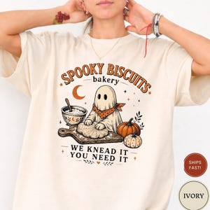 May include: An ivory t-shirt featuring a graphic of a ghost wearing a polka-dot bandana, kneading dough on a wooden board. The text reads SPOOKY BISCUITS bakery WE KNEAD IT YOU NEED IT, accompanied by a bowl, pumpkins, stars, and a crescent moon.