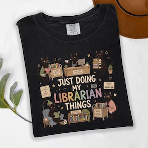 May include: A black t-shirt featuring a graphic of black cats performing library tasks. The text reads "JUST DOING MY LIBRARIAN THINGS" alongside illustrations of book carts, shelves, and library cards. The shirt has a Comfort Colors brand label.