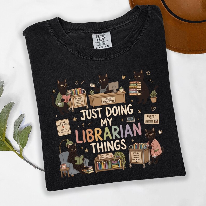 May include: A black t-shirt featuring a graphic of black cats performing library tasks. The text reads "JUST DOING MY LIBRARIAN THINGS" alongside illustrations of book carts, shelves, and library cards. The shirt has a Comfort Colors brand label.