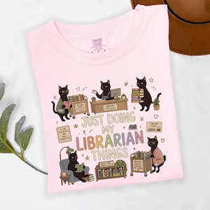Comfort Colors Blossom black cat librarian shirt featuring cute black cats reading books, library checkout desk, book cart, stacked books, library card, cozy reading chair, funny library illustrations, school librarian tee for library staff, reading teacher, book lover and BookTok fans.