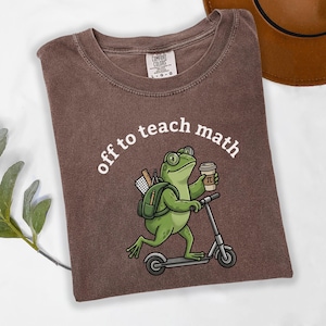 May include: A brown short-sleeve t-shirt featuring a graphic of a green frog wearing glasses, carrying a backpack with school supplies, holding a coffee cup, and riding a scooter. The text above the frog reads, off to teach math.