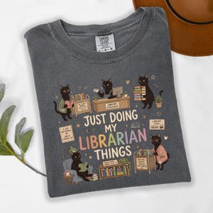 May include: A charcoal gray t-shirt featuring a graphic of black cats in a library setting. The text reads, "JUST DOING MY LIBRARIAN THINGS," surrounded by illustrations of cats reading books, a checkout desk, a book cart, and various library items.
