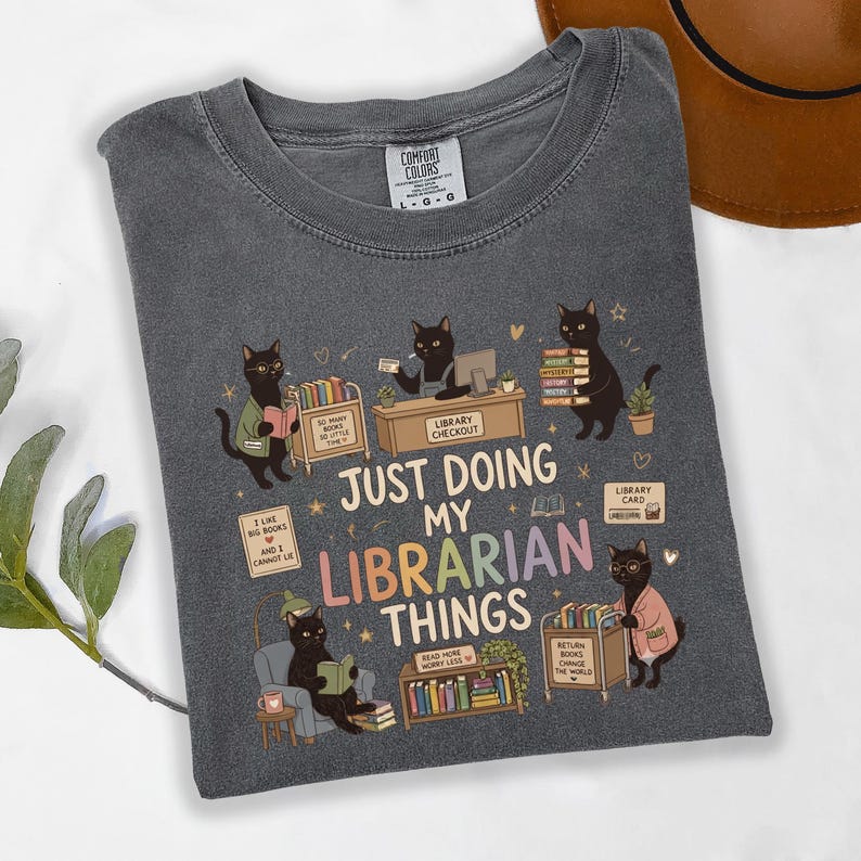May include: A charcoal gray t-shirt featuring a graphic of black cats in a library setting. The text reads, "JUST DOING MY LIBRARIAN THINGS," surrounded by illustrations of cats reading books, a checkout desk, a book cart, and various library items.