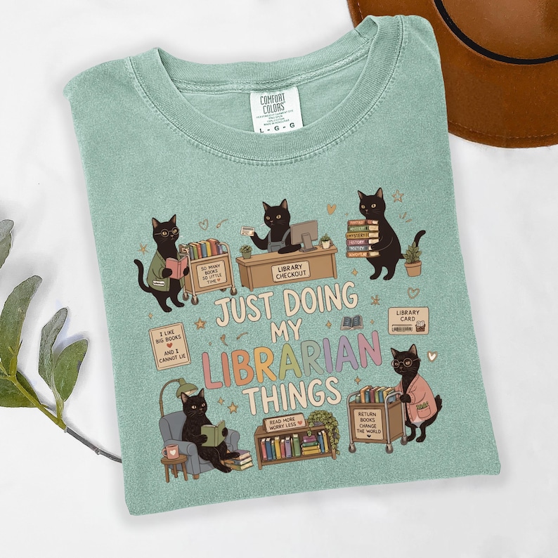 May include: A sage green t-shirt featuring a graphic of five black cats in a library setting. The text reads, "JUST DOING MY LIBRARIAN THINGS," surrounded by illustrations of book carts, a checkout desk, books, and a library card.
