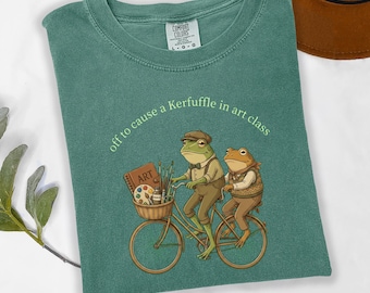 May include: A light green t-shirt featuring a graphic of two frogs riding a bicycle. One frog wears a cap and carries a basket with art supplies. The text above reads, "off to cause a Kerfuffle in art class." The shirt has a Comfort Colors label.