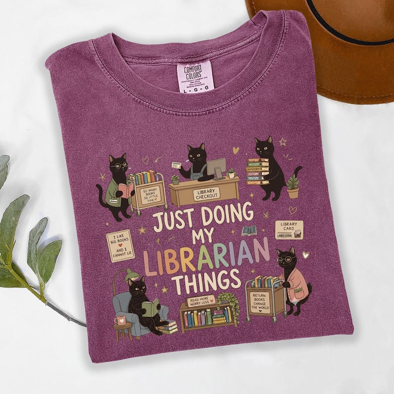 May include: A plum-colored t-shirt featuring a graphic of black cats in a library setting. The text reads: JUST DOING MY LIBRARIAN THINGS. The design includes cats reading, shelving books, and using a library cart, with various book-related icons.