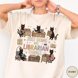 May include: An ivory t-shirt featuring a graphic of five black cats in a library setting. The text reads, "JUST DOING MY LIBRARIAN THINGS," along with smaller phrases like "SO MANY BOOKS SO LITTLE TIME" and "READ MORE WORRY LESS" among illustrations of books.