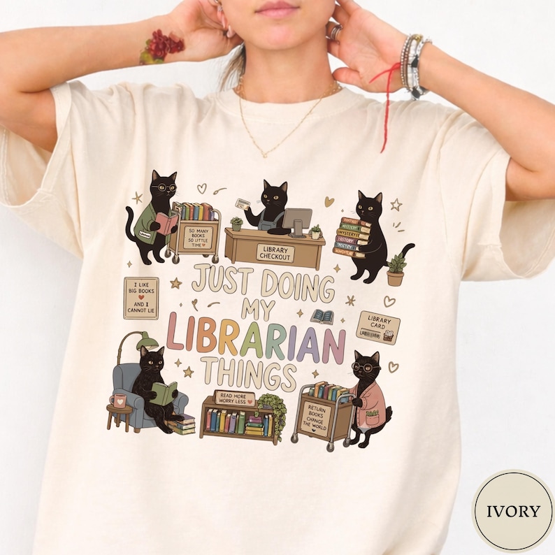 May include: An ivory t-shirt featuring a graphic of five black cats in a library setting. The text reads, "JUST DOING MY LIBRARIAN THINGS," along with smaller phrases like "SO MANY BOOKS SO LITTLE TIME" and "READ MORE WORRY LESS" among illustrations of books.