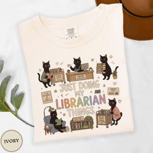 Comfort Colors Ivory black cat librarian shirt featuring cute black cats reading books, library checkout desk, book cart, stacked books, library card, cozy reading chair, funny library illustrations, school librarian tee for library staff, reading teacher, book lover and BookTok fans.
