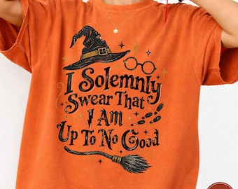 May include: An orange short-sleeve t-shirt featuring a black graphic print of a witch hat, round glasses, and a broomstick. The text reads, I Solemnly Swear That I Am Up To No Good, accompanied by small star, lightning bolt, and footprint icons.