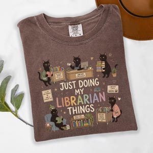 May include: A brown crew neck t-shirt featuring a graphic of black cats in a library setting. The text reads "JUST DOING MY LIBRARIAN THINGS" along with smaller phrases like "SO MANY BOOKS SO LITTLE TIME" and "I LIKE BIG BOOKS AND I CANNOT LIE."