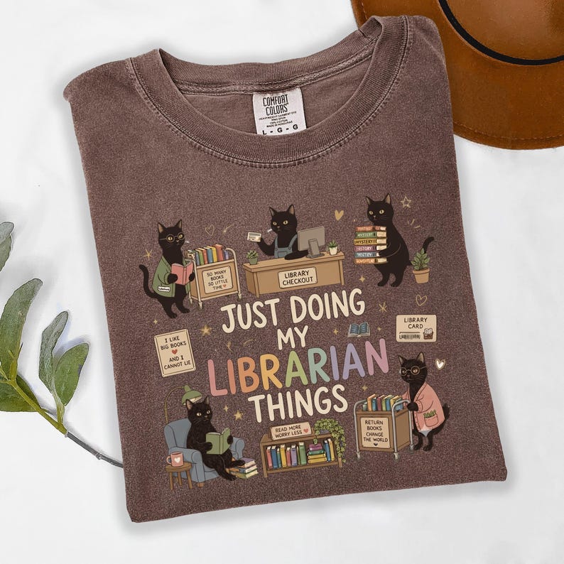 May include: A brown crew neck t-shirt featuring a graphic of black cats in a library setting. The text reads "JUST DOING MY LIBRARIAN THINGS" along with smaller phrases like "SO MANY BOOKS SO LITTLE TIME" and "I LIKE BIG BOOKS AND I CANNOT LIE."