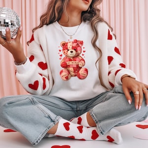 May include: White sweater with red heart accents and a teddy bear graphic. The teddy bear has a patchwork design with a heart that says "Be Mine". The sleeves and socks also have red hearts.