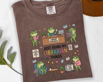May include: A brown short-sleeve t-shirt featuring a graphic of green frogs in a library setting. The text reads: "JUST DOING MY LIBRARIAN THINGS," "LIBRARY CHECKOUT," "I LIKE BIG BOOKS AND I CANNOT LIE," "READ MORE WORRY LESS," and "RETURN BOOKS CHANGE THE WORLD."