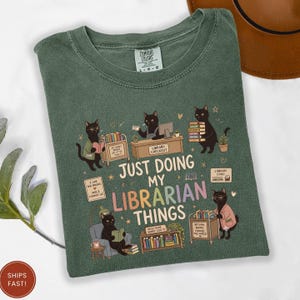 May include: A sage green t-shirt featuring a graphic of black cats performing library tasks. The text reads, "JUST DOING MY LIBRARIAN THINGS." The design includes cats reading, shelving books, and working at a checkout desk, surrounded by small stars and hearts.
