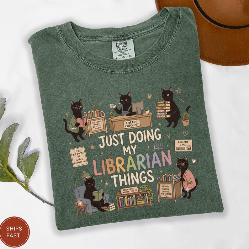 May include: A sage green t-shirt featuring a graphic of black cats performing library tasks. The text reads, "JUST DOING MY LIBRARIAN THINGS." The design includes cats reading, shelving books, and working at a checkout desk, surrounded by small stars and hearts.