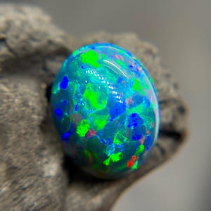 May include: A polished, oval-shaped opal gemstone. The opal displays a vibrant play-of-color with flashes of green, blue, and red hues. The gemstone is set against a dark, textured background, highlighting its iridescent qualities.