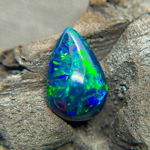 May include: A teardrop-shaped black opal gemstone with vibrant internal colors of blue, green, and red. The polished surface reflects light, showcasing the play-of-color effect. The gemstone is set against a dark, textured background.