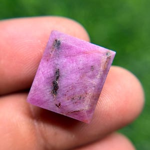 May include: A square-cut, translucent pink gemstone with a faceted surface. The stone displays a vibrant magenta hue with visible natural inclusions, including dark, linear mineral streaks running vertically across the center of the face.