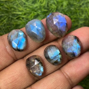 Puede incluir: Seis piedras preciosas de labradorita facetadas con tonos azules y grises. Las piedras son de forma ovalada y tienen una superficie pulida y reflectante. Las piedras se sostienen en una mano sobre un fondo verde.