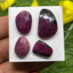 Może przedstawiać: Cztery fasetowane, ciemnoczerwone kamienie szlachetne rubinu o różnych kształtach są prezentowane na białej powierzchni. Kamienie mają wypolerowaną, odblaskową powierzchnię, z niektórymi ciemnymi wtrąceniami. Tło jest rozmyte, z nutami zieleni i żółci.