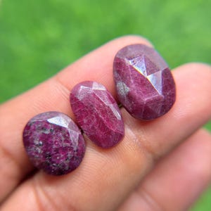 Può includere: Tre rubini sfaccettati di forma ovale in tonalità magenta intenso e rosso scuro. Le pietre presentano una superficie lucida e riflettente con inclusioni interne visibili e texture cristalline naturali.