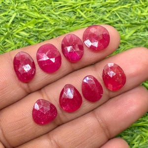 Puede incluir: Una colección de rubíes facetados de forma ovalada y de lágrima. Las piedras preciosas son de un color rojo intenso y vibrante, presentadas sobre un fondo de hierba verde. Medidas en centímetros.