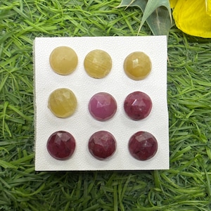 Puede incluir: Nueve gemas redondas en tonos amarillos y rojo-púrpura están dispuestas sobre una tarjeta blanca. Las gemas tienen una superficie facetada. El fondo es verde, con algunos pétalos de flores amarillas visibles.