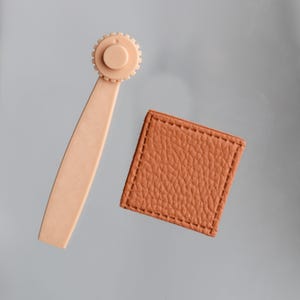 Peut inclure: Un cutter rotatif beige clair avec une tête en forme de roue dentée et une longue poignée, à côté d'un patch carré en cuir marron texturé avec des bords cousus. Les articles sont sur fond gris.