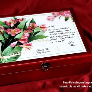 May include: A red mahogany keepsake box with a hinged lid. The lid features a custom printed ceramic tile with a bouquet of pink flowers and a personalised message that reads "Happy 75th July 28, 2021" and "Cousins are a little bit of childhood that was never to be lost. And only a few of them are very lucky to have grown to be best friends. So grateful to have you in my life. Love, Sharon."