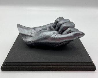 May include: A silver-colored hand sculpture, resting on a black rectangular base. The hand is in a cupped position, with fingers slightly curled. The sculpture has a metallic sheen, and the base has a textured surface.