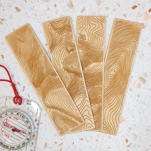 May include: Four wooden bookmarks featuring a topographic map design. The bookmarks are made from light brown wood and have a natural finish.